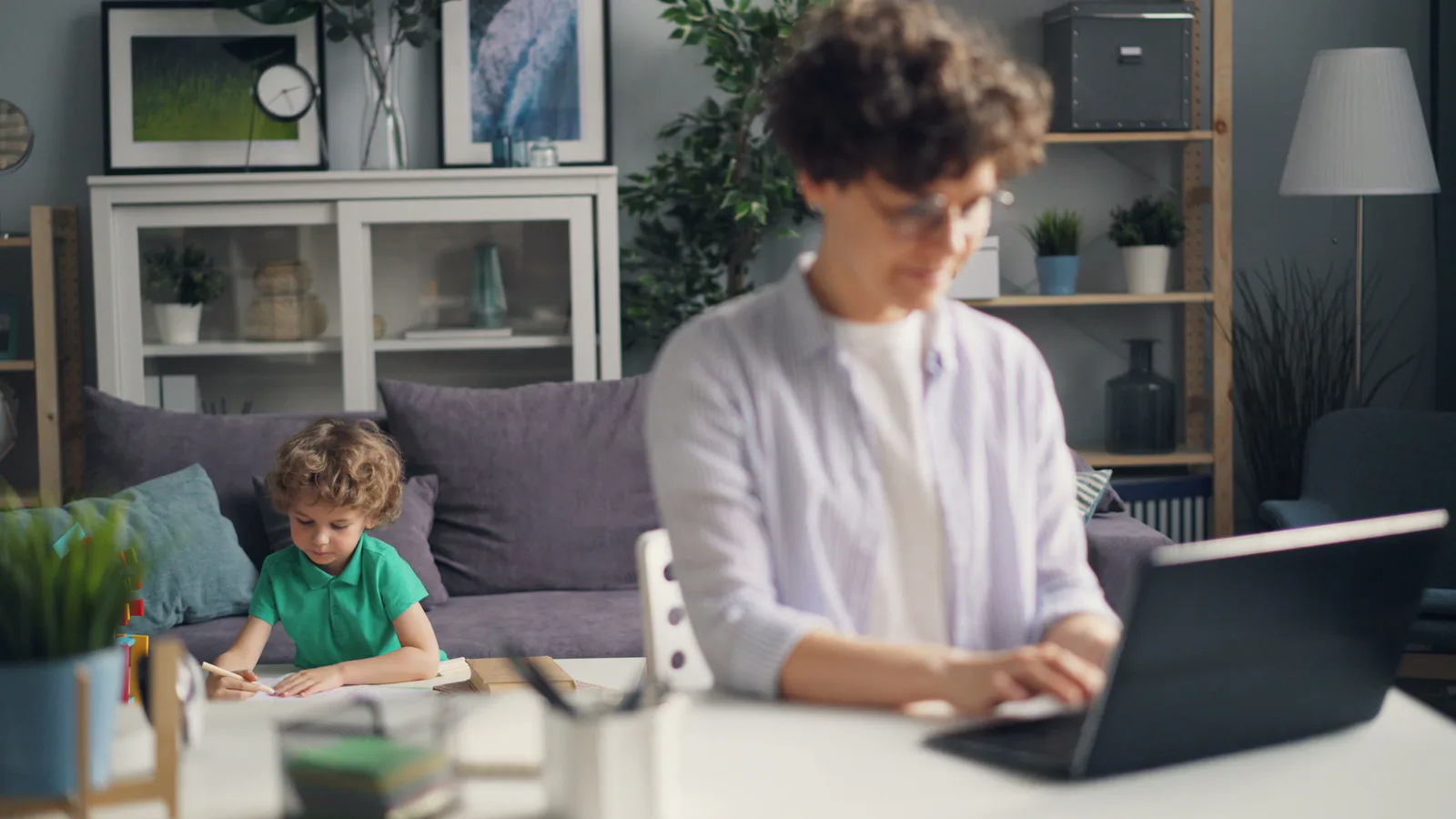 Parent using a laptop while a child studies nearby at home